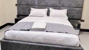 A modern bedroom with a large bed, grey upholstered headboard, white bedding, and a bedside table with a lamp. - Lee Villas  (Kampala)