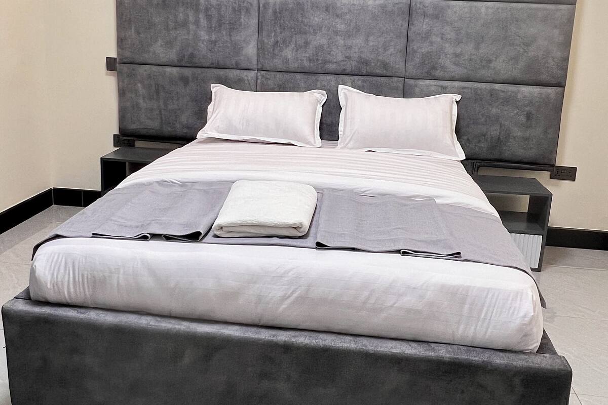 A modern bedroom with a large bed, grey upholstered headboard, white bedding, and a bedside table with a lamp.