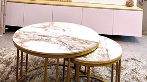 A modern living room with a large flat-screen TV mounted on a white wall with a marble design. There is a wooden panel and a white cabinet below the TV. In front of the TV, there is a round marble table with two chairs. - Lee Villas  (Kampala)