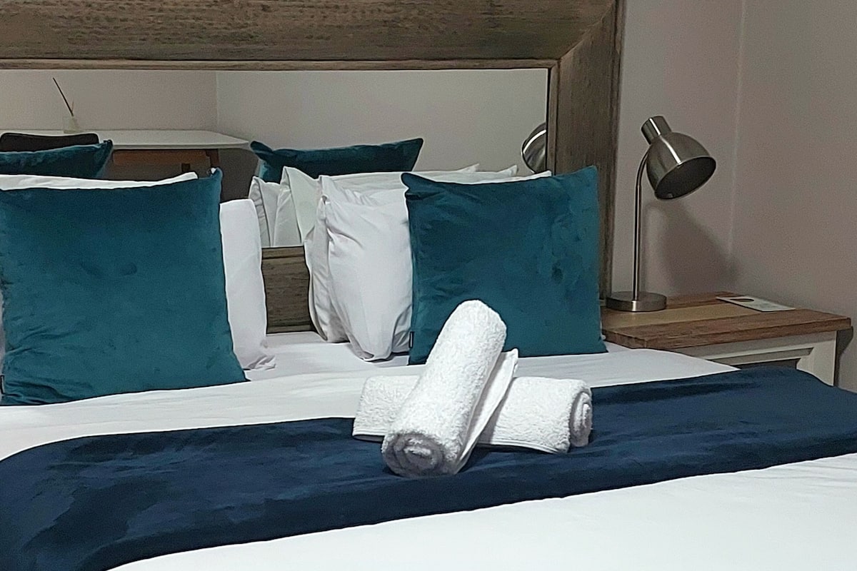 Une chambre d’hôtel avec un lit bien fait, une tête de lit en bois, une table de chevet avec une lampe, et une décoration aux couleurs bleu et blanc.