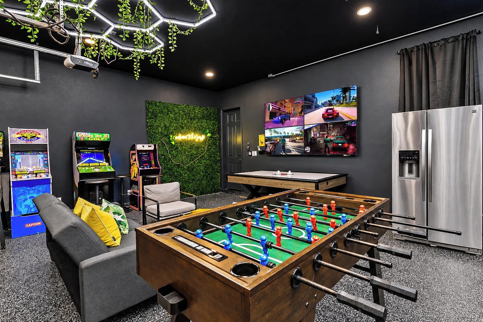 Una stanza con un tavolo da calcio, giochi arcade, un divano, una TV a parete e un frigorifero.