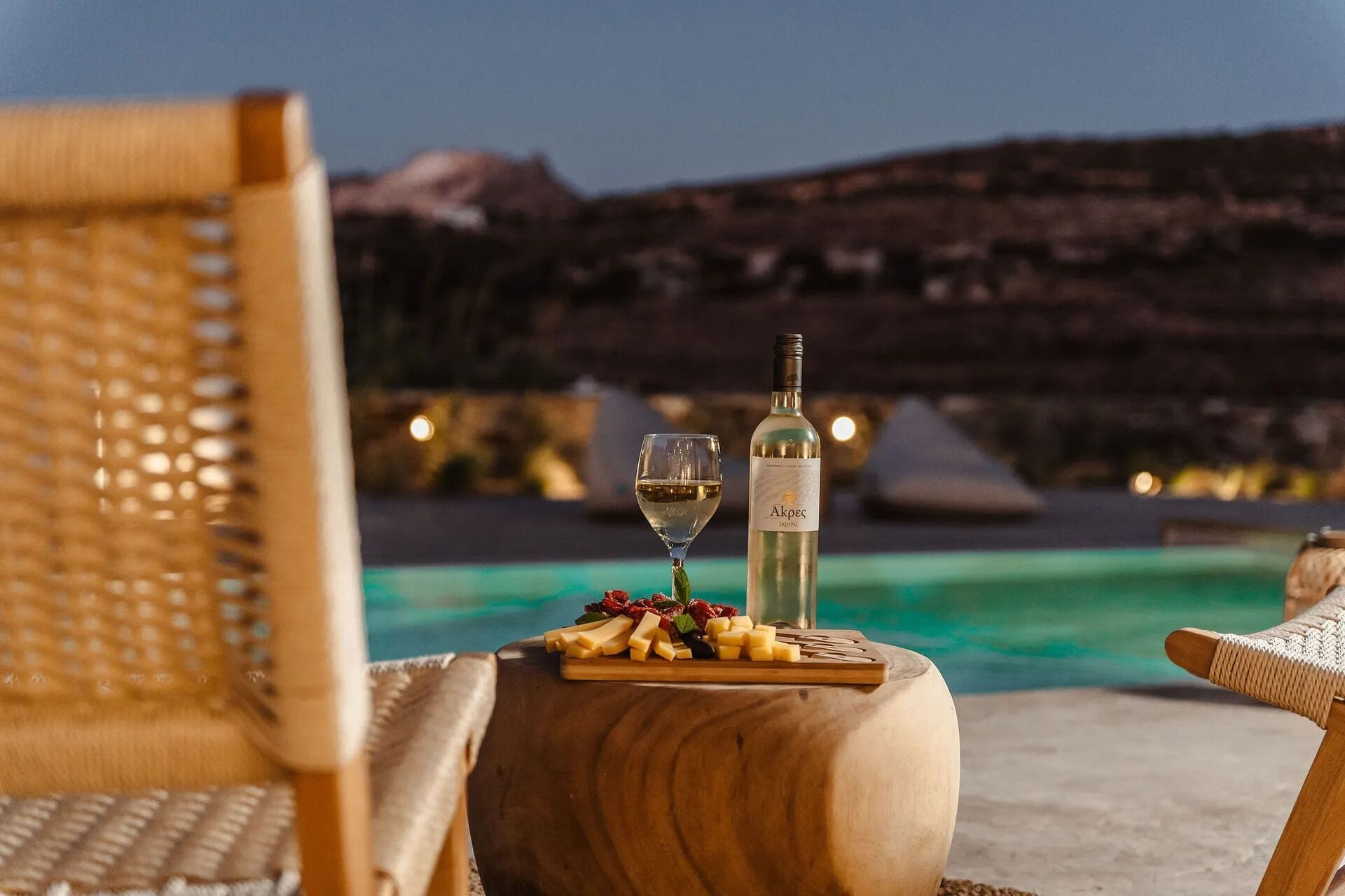 Una mesa de madera con una botella de vino Akpec, una copa de vino blanco y una tabla de quesos junto a una piscina con montañas al fondo.