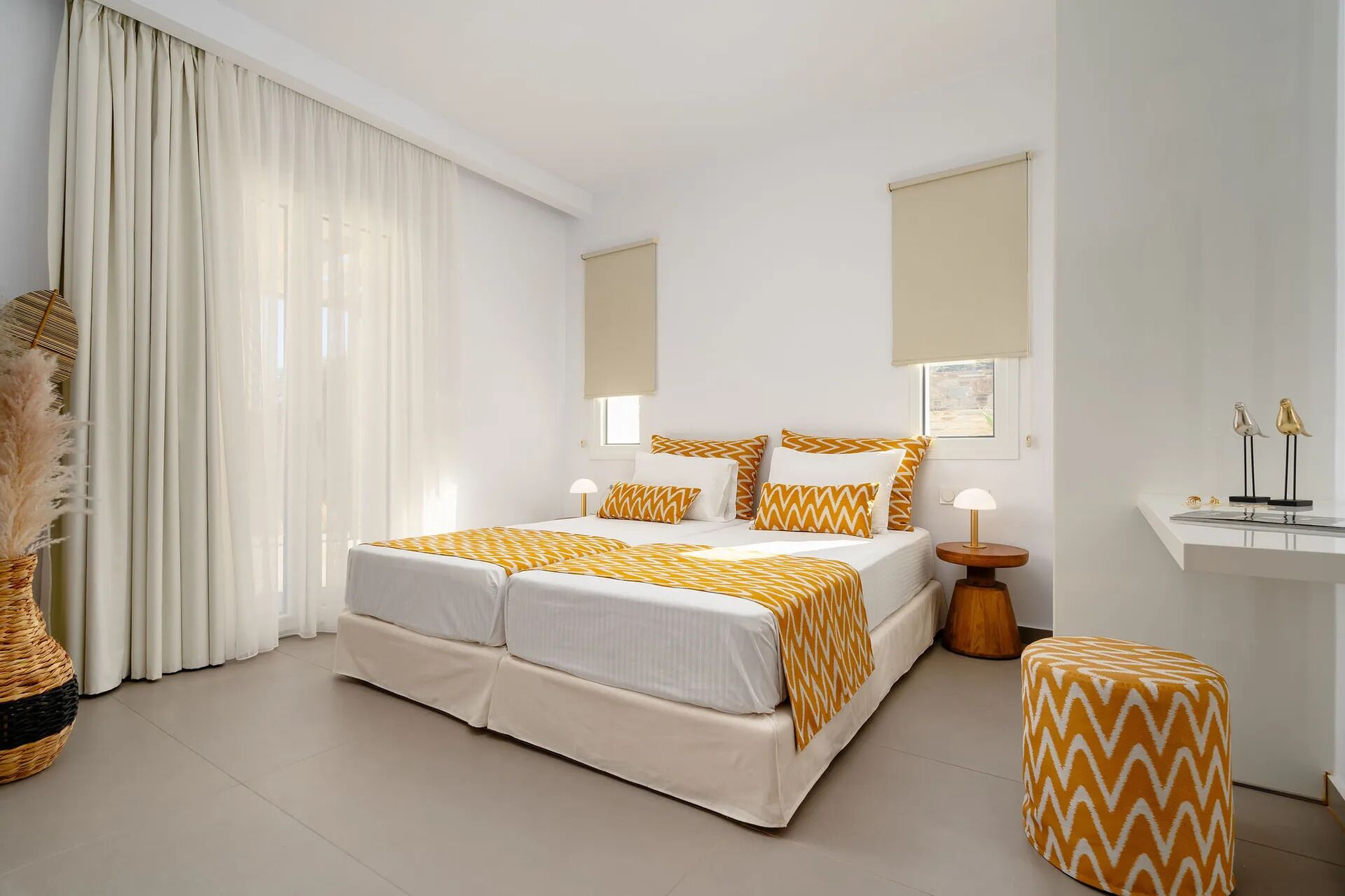 A hotel room with a bed, two bedside tables, a nightstand, a window with curtains, a wall-mounted mirror, and a small shelf with decorative items.