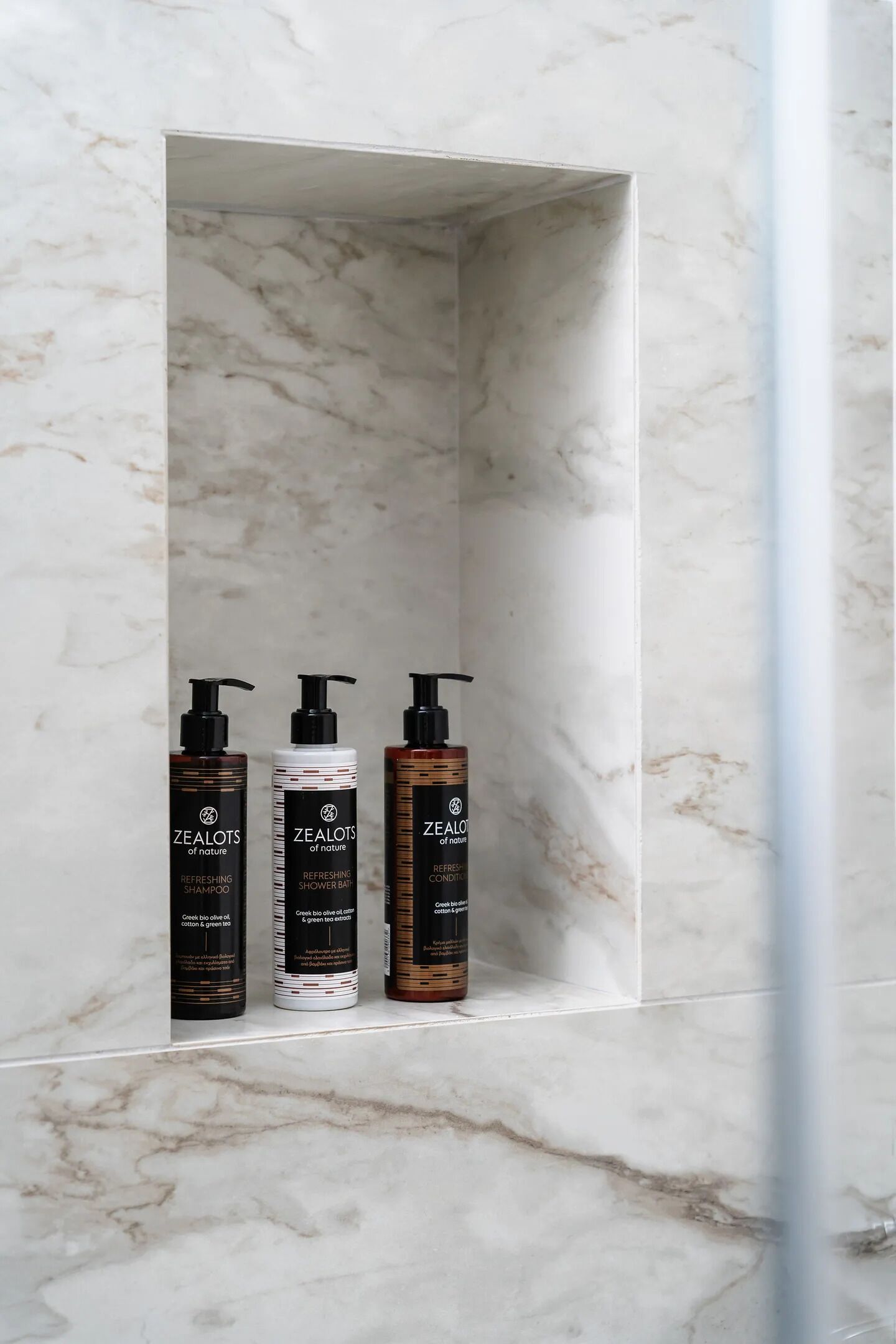 A bathroom with a marble wall and a shelf containing three bottles of Zealots of Nature shampoo and conditioner.