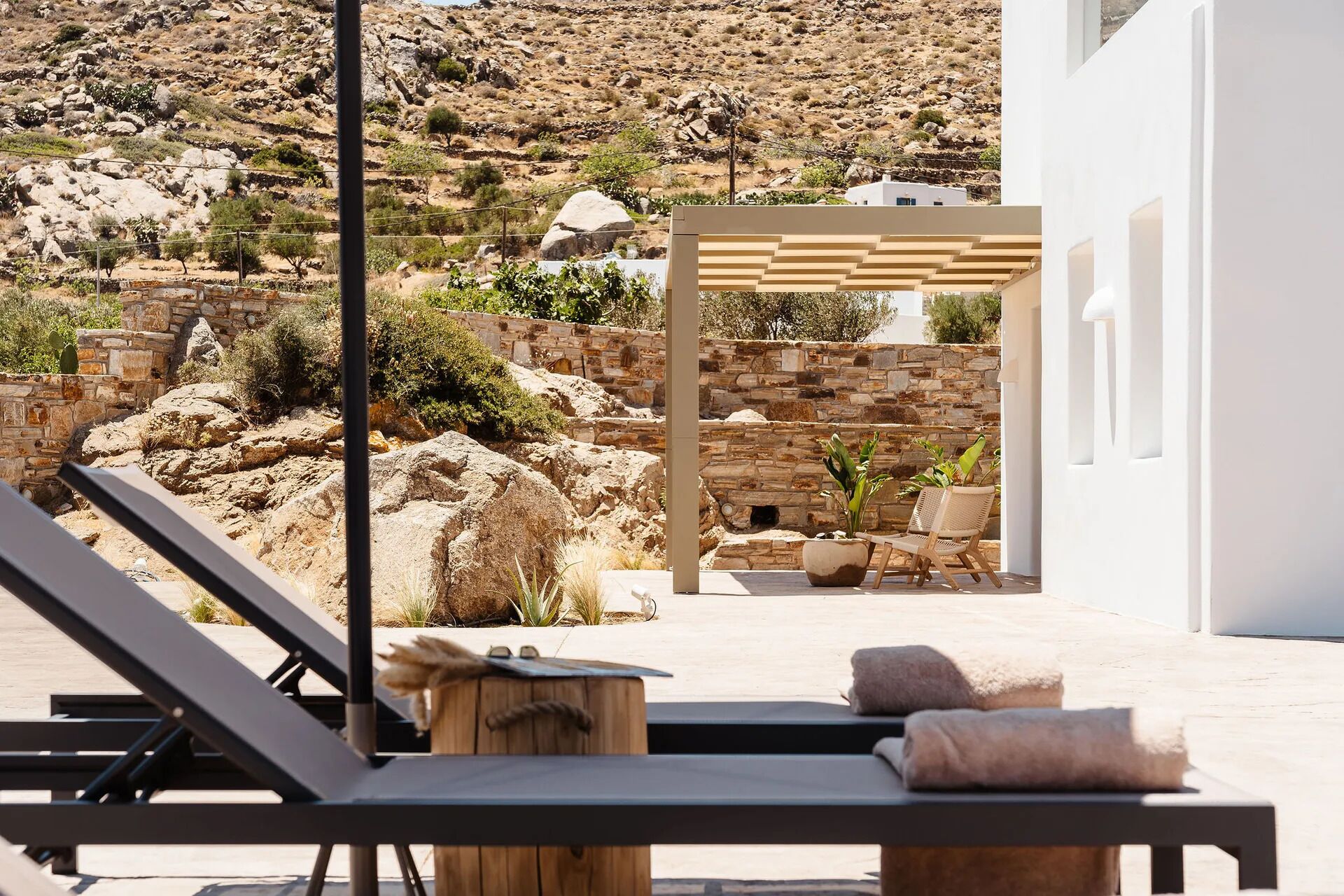 Un espace de détente extérieur baigné de lumière, avec un mur en pierre, une pergola en bois et une vue sur une colline rocheuse.
