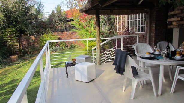 Un patio con mesa y sillas, una mesita con una botella y un vaso, y vistas a un jardín con muro de ladrillo y árboles.
