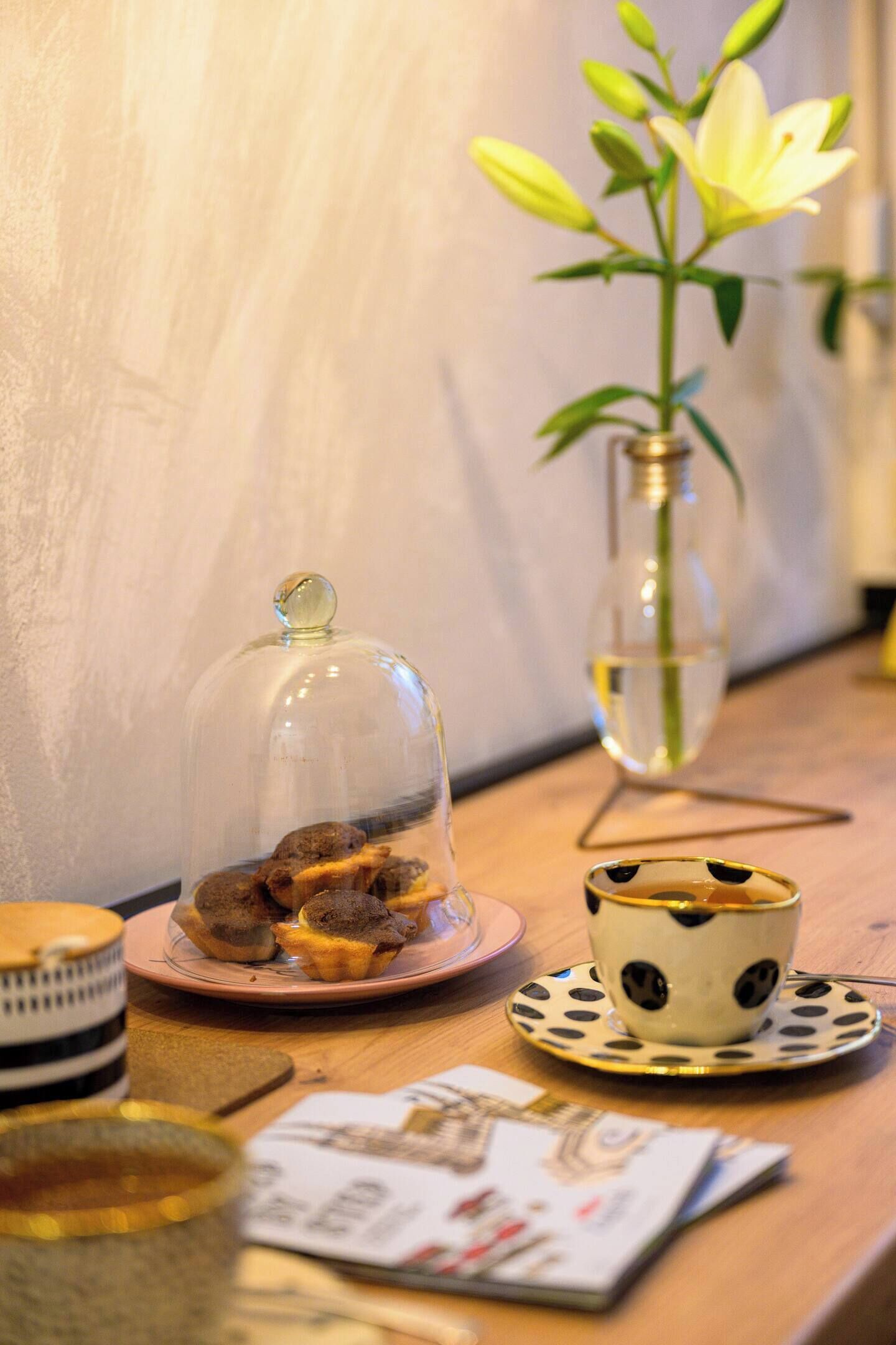 Una mesa de madera con una campana de vidrio, una taza de lunares, un florero con una flor y algunos papeles.