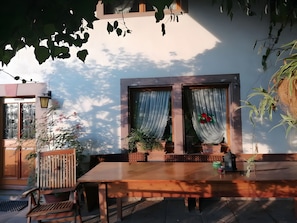 Un juego de mesa y banco de madera frente a un edificio con dos ventanas, una con cortina y otra adornada con flores.