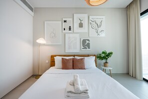A neatly made bed with white linens and a folded towel, a bedside lamp, a small table with a potted plant, and several framed artworks on the wall.