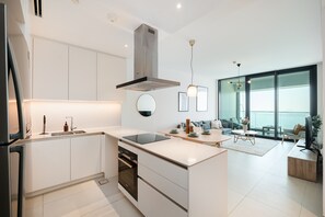 A modern kitchen with white cabinetry, a built-in oven, and a central island. There is a dining area with a round table and chairs, and a large window offering a view of the outdoors.