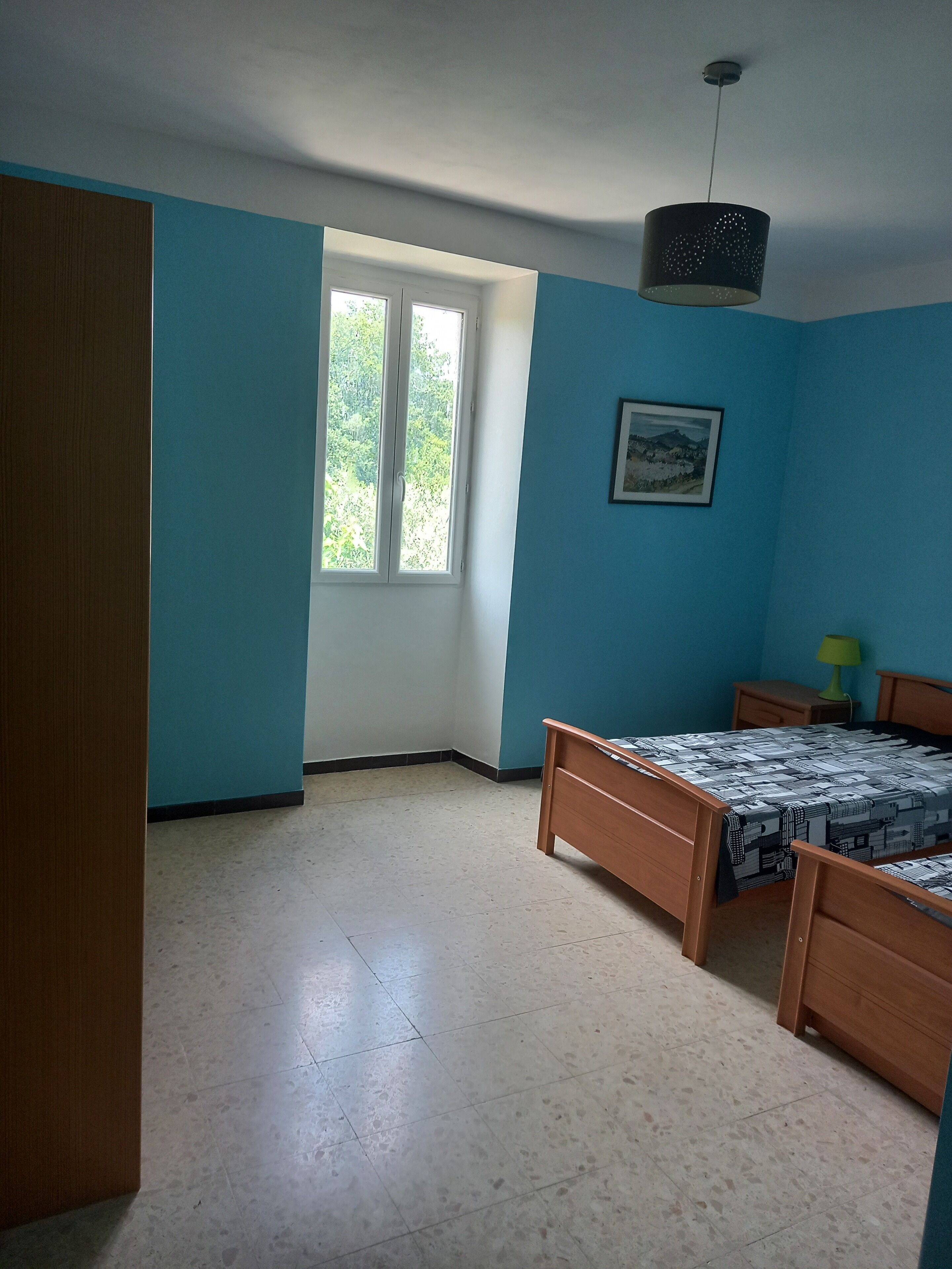 Une chambre à coucher avec deux lits en bois, une fenêtre donnant sur un paysage verdoyant, un tableau encadré au mur et un luminaire au plafond.