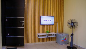 A room with a black door, a television mounted on the wall, a small shelf with a plant, a refrigerator, and a standing fan. - BON HOMES (Ibadan)