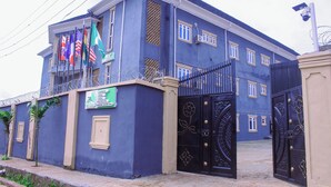 A large, two-story building with a blue facade, a gated entrance, and multiple flags. - BON HOMES (Ibadan)