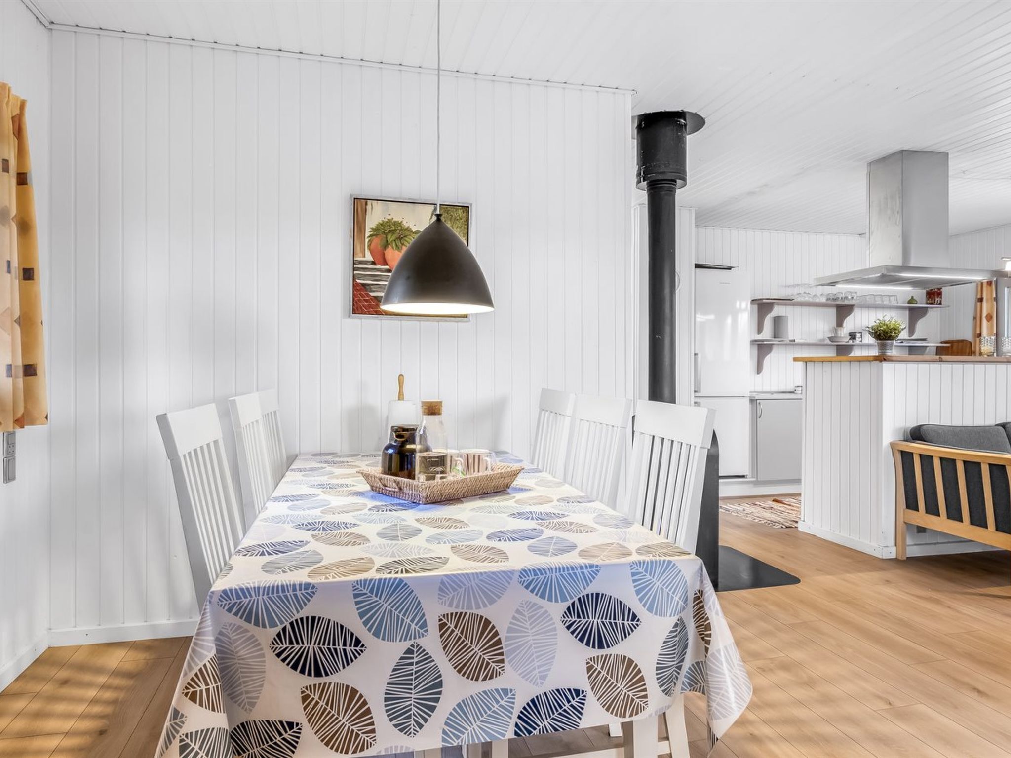 Un coin repas avec une table recouverte d’une nappe à motifs de feuilles, des chaises blanches, un luminaire suspendu et une cuisine en arrière-plan.