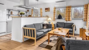 Una sala de estar moderna y bien iluminada con un sofá gris, una mesa de centro de madera y una mesita auxiliar. Se ve una zona de cocina con armarios blancos y una ventana con cortinas al fondo.