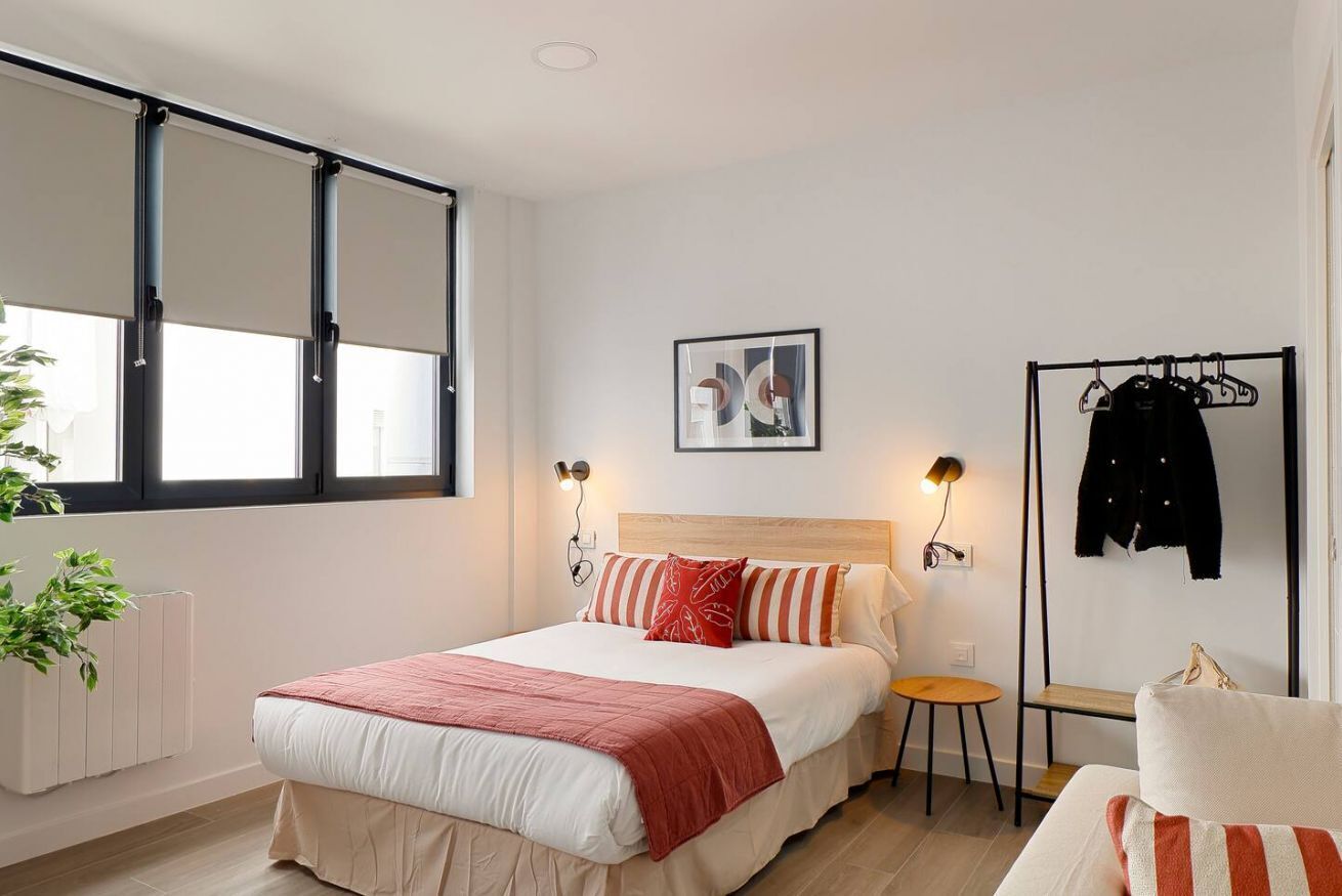 Un dormitorio con cama, mesitas de noche, radiador, ventana con persianas, perchero y un cuadro en la pared.