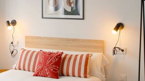 Un lit avec une tête de lit en bois, des oreillers à rayures et un oreiller rouge orné d’un motif blanc. Deux lampes murales sont fixées de part et d’autre du lit.