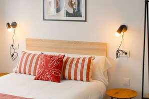 Ein Bett mit einem Holz-Kopfteil, gestreifte Kissen und ein rotes Kissen mit einem weißen Muster. An beiden Seiten des Bettes befinden sich zwei Wandlampen.
