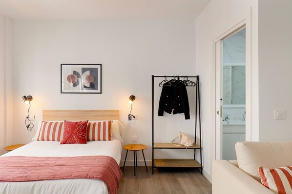 A bedroom with a bed, bedside tables, a coat rack, a chair, and a bathroom visible through an open door.