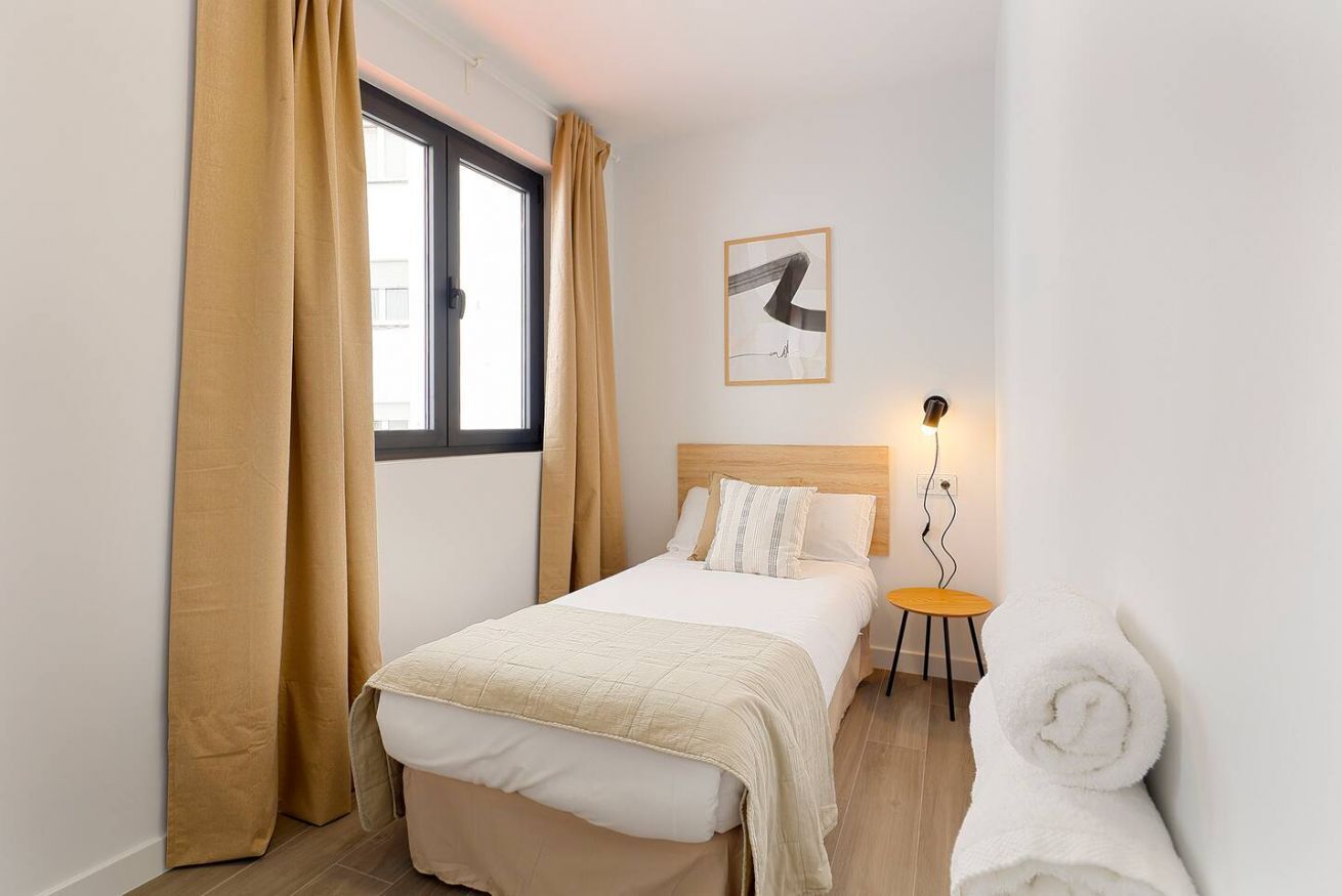 Quarto de hotel com uma cama, uma mesinha com uma luminária, uma toalha enrolada e uma janela com cortinas.