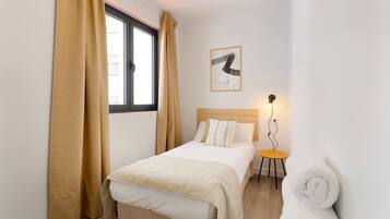 Quarto de hotel com uma cama, uma mesinha com uma luminária, uma toalha enrolada e uma janela com cortinas.