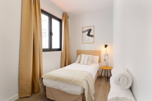 A hotel room with a bed, a small table with a lamp, a rolled towel, and a window with curtains.