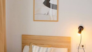 Een slaapkamer met een houten hoofdbord, een bed met witte en gemêleerde kussens, een lijstwerk abstract kunstwerk en een wandlamp.