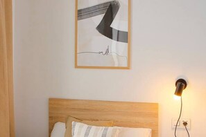 Un dormitorio con cabecera de madera, cama con almohadas blancas y estampadas, una obra de arte abstracta enmarcada y una lámpara fijada a la pared.