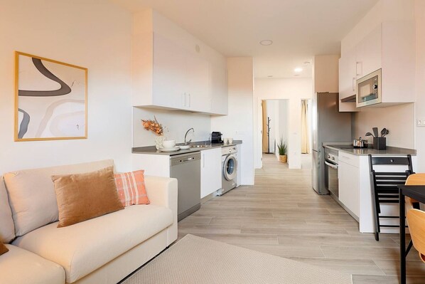 A modern kitchen with a dishwasher, washing machine, and microwave. There is a sofa with cushions and a dining area with a table and chairs.