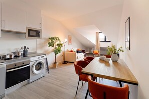 Une cuisine moderne dotée d’un four et d’un micro-ondes encastrés, un coin repas avec une table en bois et des chaises orange, et un espace de vie comprenant un canapé et une plante.