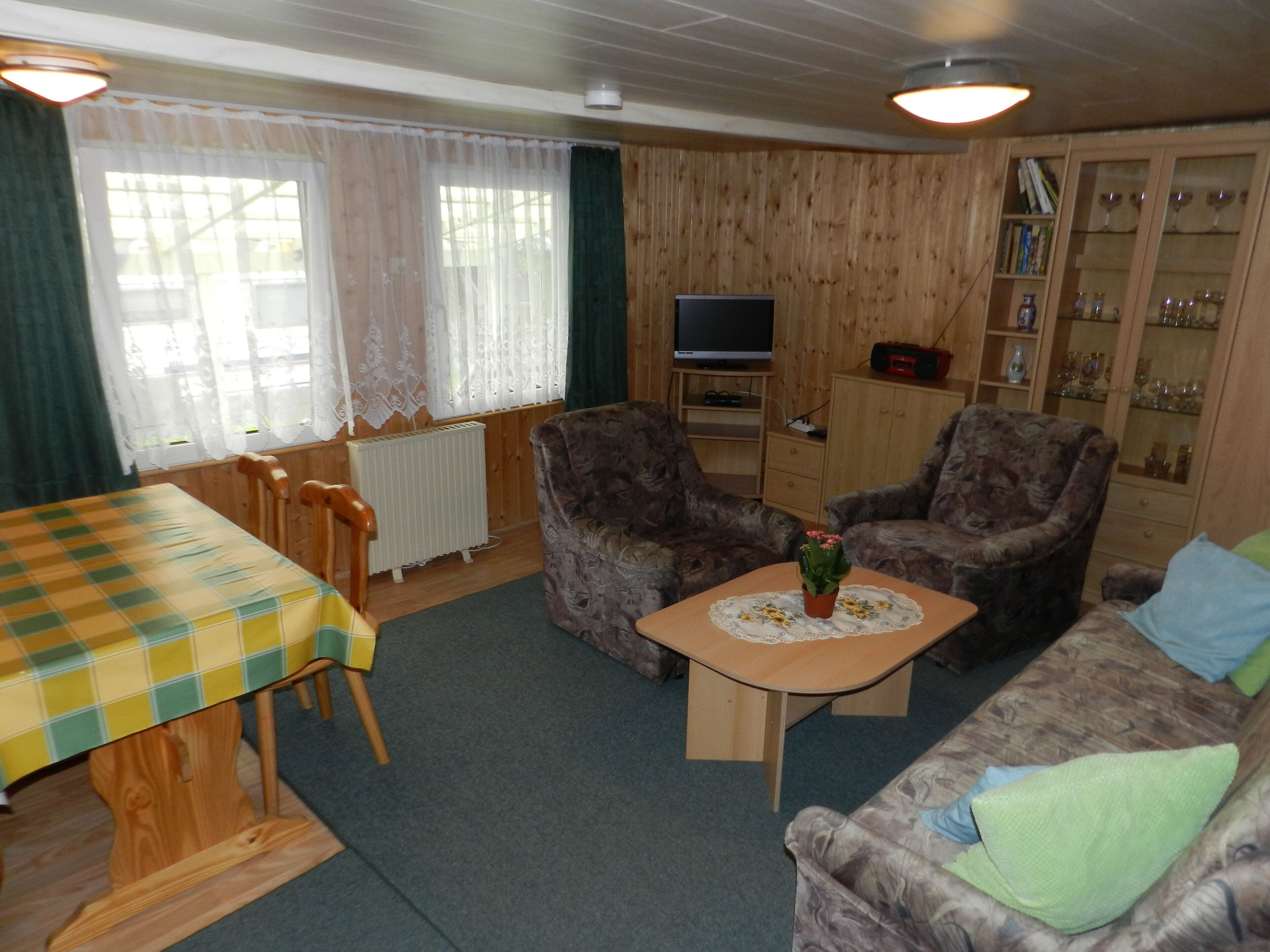 A cozy living room with a wooden floor, a dining table with a checkered tablecloth, two armchairs, a sofa, a television, and a cabinet with glassware.