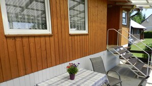 Pared exterior de madera con una ventana, una mesa cubierta con mantel escocés y asientos al aire libre.