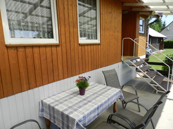 A wooden exterior wall with a window, a table with a plaid tablecloth, and outdoor seating.