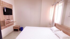 A bedroom with a bed, a TV mounted on the wall, a small wooden nightstand, and a blue trash bin. - Hotel Rewa Regency (Huzur)