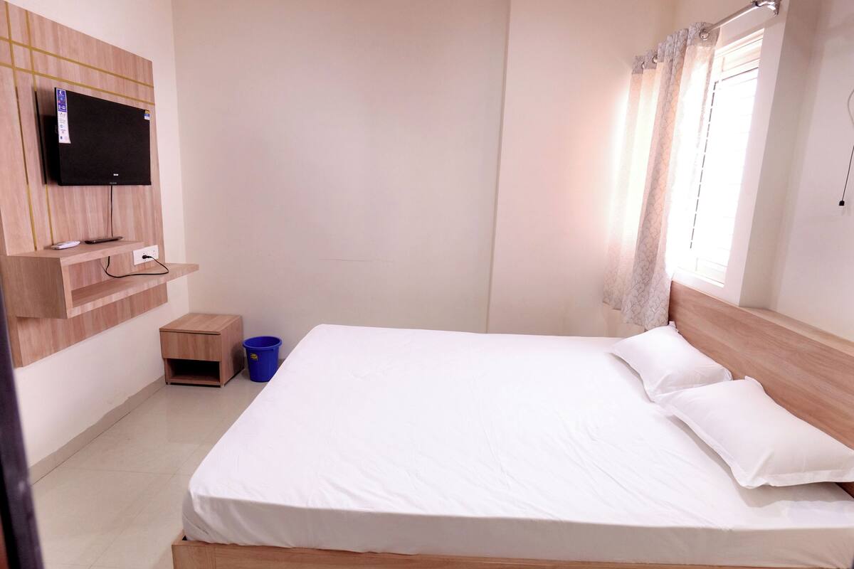 A bedroom with a bed, a TV mounted on the wall, a small wooden nightstand, and a blue trash bin.