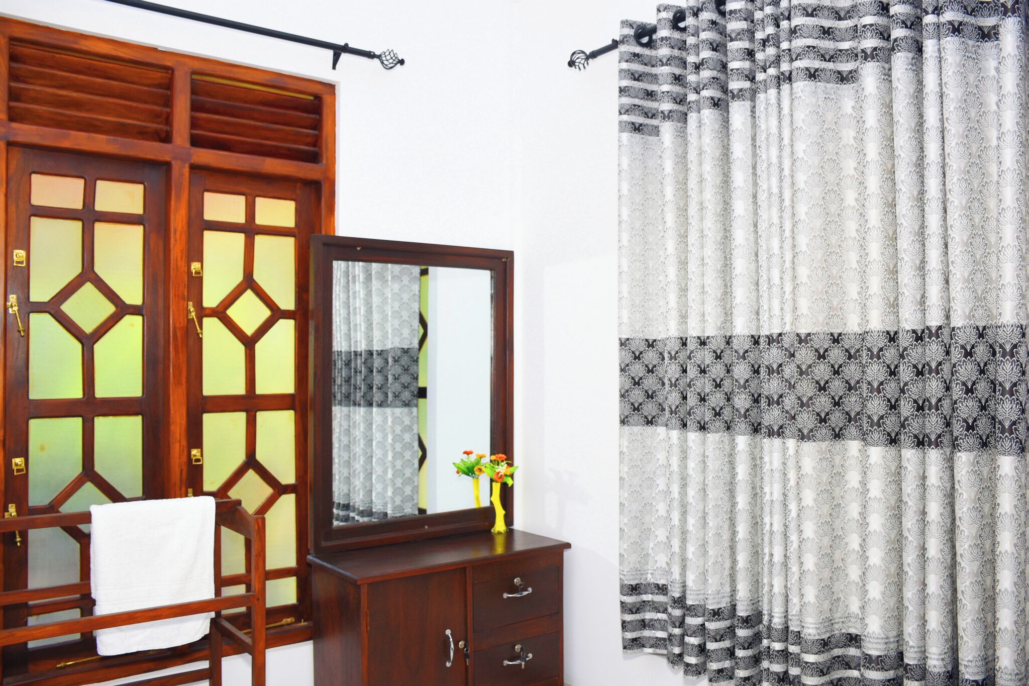 A room with a wooden wardrobe, a mirror, a small cabinet, and a patterned curtain.