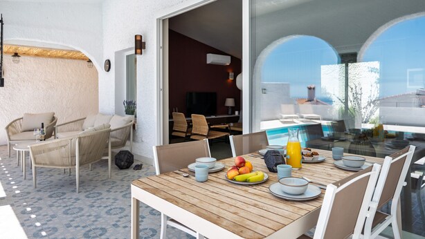 A dining area with a wooden table, white chairs, and a glass partition. There are fruits and a bottle on the table.
