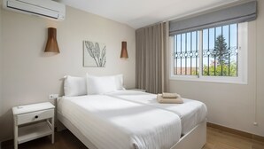 A bedroom with a bed, bedside table, wall-mounted lamp, air conditioning unit, and a window with curtains.