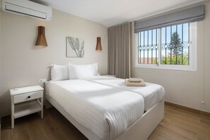 A bedroom with a bed, bedside table, wall-mounted lamp, air conditioning unit, and a window with curtains. - Villa Los Pomelos - Capistrano! (Nerja)