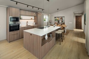 A modern kitchen with a central island, built-in appliances, and a dining area with a wooden table and chairs. - Villa Los Pomelos - Capistrano! (Nerja)