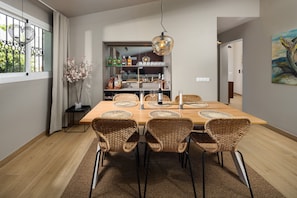 A dining area with a wooden table, wicker chairs, a hanging light fixture, and a shelf with bottles and glasses. - Villa Los Pomelos - Capistrano! (Nerja)