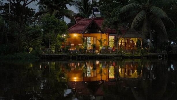 A house with a red roof, surrounded by water and palm trees, reflecting the structure and lights.