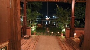 An outdoor wooden deck with a thatched roof, surrounded by palm trees and a pathway leading to a distant view.