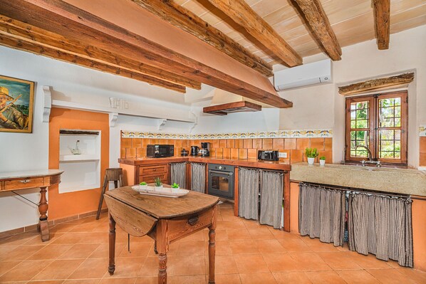 Une cuisine avec des poutres en bois, un sol carrelé, une table en bois et une fenêtre avec des rideaux.
