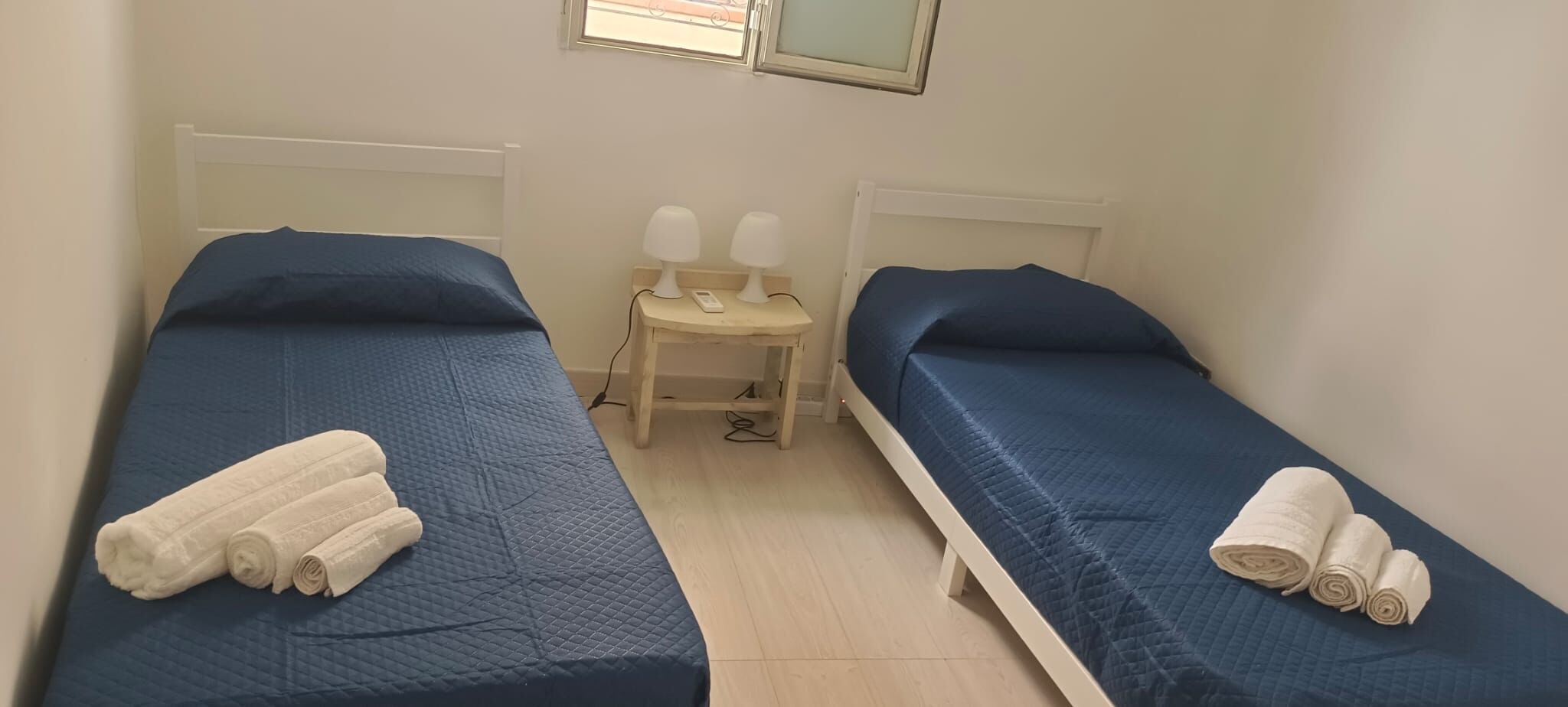 Une petite chambre avec deux lits simples, chacun recouvert d’une couverture bleue et agrémenté de serviettes blanches. Une petite table en bois, sur laquelle sont posées deux lampes blanches, se trouve entre les lits.