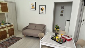 Un petit espace de vie accueillant, comprenant un canapé, une table à manger avec des chaises, un coin cuisine avec un évier et des placards, et une porte donnant sur une autre pièce.
