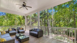 A covered outdoor area with wicker furniture, a ceiling fan, and large windows offering a view of a wooded area. - Seaclusion at the Beach (Pawleys Island)