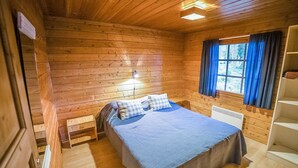 Una habitación de cabaña de madera con cama, mesita de noche, ventana con cortinas y un radiador.
