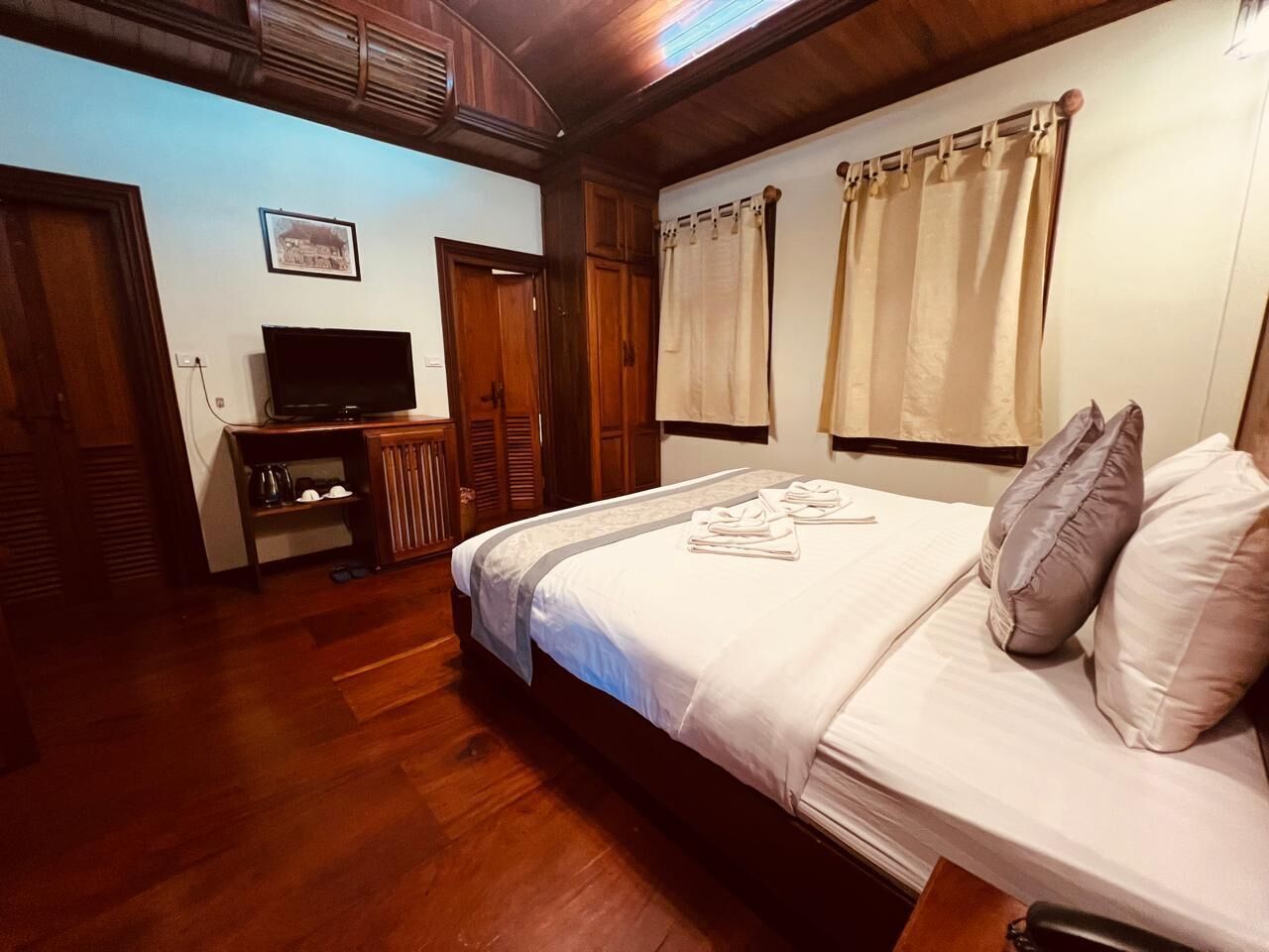 A hotel room with a wooden floor, a bed with white bedding, a television on a wooden stand, and a window with curtains.