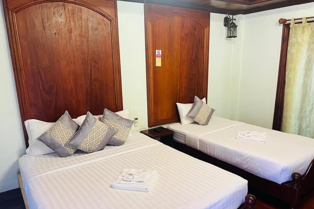 A hotel room with two beds, each with a wooden headboard, white linens, and patterned pillows. There is a nightstand between the beds, and a small table with a lamp.